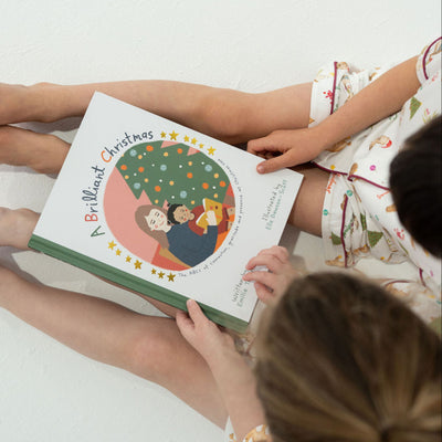 Two children sitting on a white floor holding a book titled 'A Brilliant Christmas'.