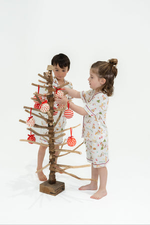Two children in pajamas decorating a small Christmas tree with red ornaments on a white background.