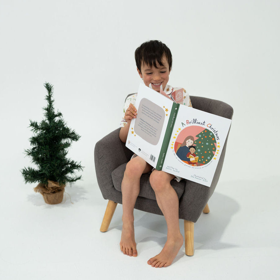 Child sitting in a chair reading a book with a small Christmas tree in the background